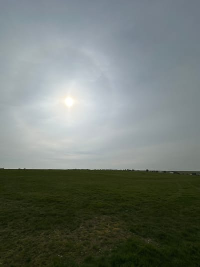 The image displays an expansive, open landscape under an overcast sky near Amesbury, United Kingdom. The upper 60% of the frame is a light grey, cloudy sky. The sun, appearing as a brilliant white disc with a distinct halo, is positioned in the upper-left quadrant, indicating a hazy or diffused light source. The lower 40% of the frame is a vast, flat field of short, green grass, extending from the foreground to a distant, flat horizon. Along the horizon line, faint silhouettes are discernible: on the far left, a distinct arrangement of vertical megaliths consistent with Stonehenge is partially visible. To the right along the horizon, scattered low-rise structures and tree lines are present. No individuals, animals, vehicles, or explicit actions are observed within the scene.