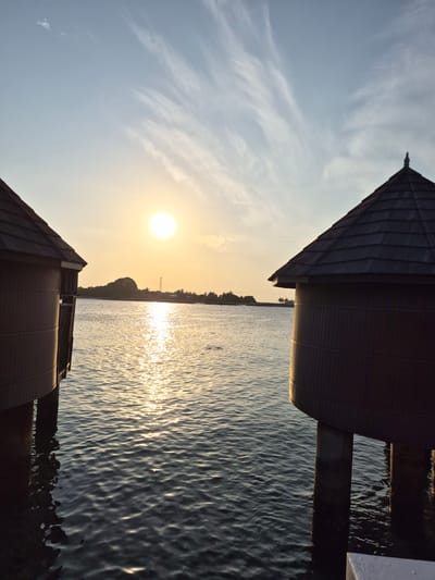 The image depicts a serene scene at Bodufolhudhoo, Maldives, at sunset. The sun is positioned centrally in the frame, casting a golden glow across the water. Reflections of the sun shimmer on the water surface, creating a visually striking pattern. Two structures with pointed roofs are visible, framing the scene on either side. The structures appear to be situated on the water's edge, possibly piers or small buildings extending into the water. The sky above the sun is clear with some whispy clouds. In the distance, a strip of land is visible, silhouetted against the bright sunlight.