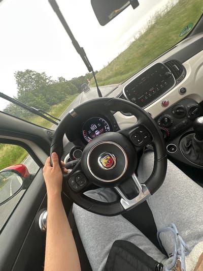 The image shows the interior of a car, an Abarth, with a driver holding the steering wheel. The car is moving, as indicated by the speedometer showing 68 km/h and the blurry background visible through the windshield. The car is traveling on a road with trees and vegetation on either side. Based on the settings in the infotainment system, one channel is titled “ANTTHUE Bay2”, which might be referring to a radio station. The exact location in Rotenburg an der Fulda, Germany, cannot be determined from the image.