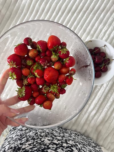 A person's hand, with a manicured nail, is holding a clear glass bowl filled with fresh strawberries. The bowl contains a large quantity of bright red strawberries, some with green stems and leaves, and a few are slightly lighter in color, indicating ripeness. The glass bowl is slightly damp, suggesting the strawberries may have been recently washed. To the right of the bowl, a white ceramic dish holds a pile of dark red cherries, each with a stem. The background consists of a textured white surface, likely a blanket or upholstery, with ribbed lines running diagonally. The lower portion of the image shows a person's lap covered by black and white patterned fabric, possibly clothing. The scene appears to be indoors, on a comfortable surface, with a focus on the fresh fruit. There is no indication of the city of Ivatsevichy, Belarus in this image.