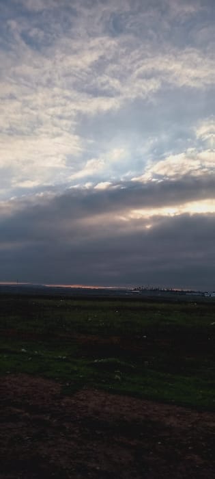 A landscape shows a bright, cloudy sky transitioning to a darkening land. The sky consists of light and dark gray clouds, with a bright area suggesting sunlight piercing through. The land is divided, with one side appearing green and vegetated, gradually darkening into brown on the other. The division between the sky and land is slightly blurred, suggesting a distant horizon or atmospheric effect.