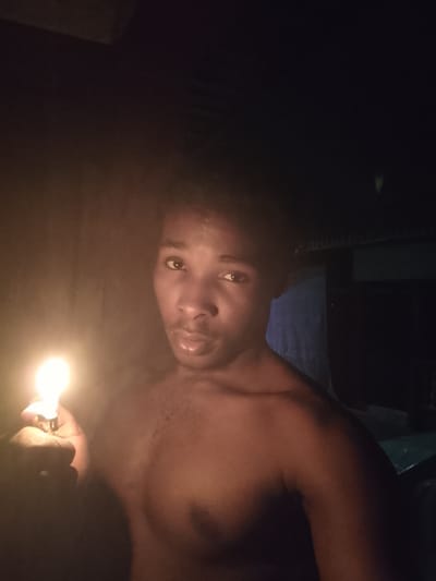 A medium shot shows a young Black male holding a lit lighter in his left hand in the foreground. He is looking directly at the camera with a neutral expression. The lighter is illuminating his face and chest, creating a warm glow. The background is dark and out of focus, with some hints of what appears to be a corrugated metal roof structure and a blue tarp or cloth. The overall lighting suggests the image was taken in low-light conditions, possibly at night, in a rural or informal setting in Orodo, Nigeria. The man is shirtless, and his upper body is visible. The composition is tightly framed, focusing on the subject and the light source.