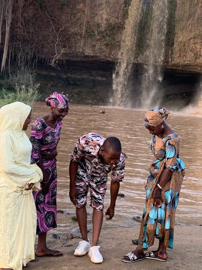Four individuals stand near a waterfall at Gurara Waterfall in the city of Kukum, Nigeria. One woman is dressed in a blue and brown patterned robe and headwrap. Another woman wears a black and pink patterned robe and headwrap. A third woman wears a long, pale yellow robe with a hood. A man stands between the two women, wearing a tan, brown, and black patterned shirt and shorts. The waterfall is on the left of the frame. The ground is covered in mud and debris.