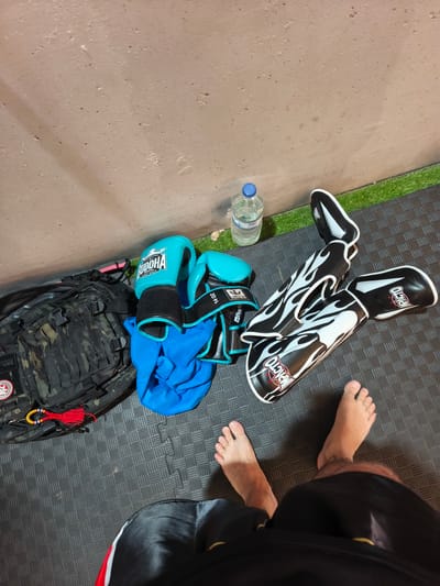 The image shows a person standing on a black interlocking foam mat in Polígon Industrial De Picassent, Spain. The person is barefoot, wearing black and red shorts. To the left of the person, there is a camouflage backpack, a pair of light blue "BUDDHA" boxing gloves, and a blue towel. Near the gloves is a bottle of water. To the right of the gloves, there is a pair of black and white shin guards that say "IMPACTO." The background includes a beige wall and a strip of artificial green grass at the bottom of the wall.
