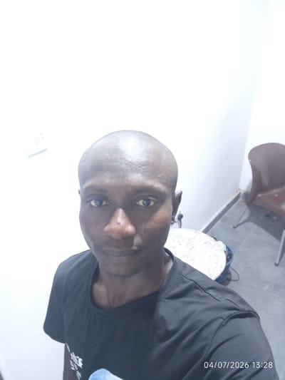 A bald Black man is taking a selfie. He is wearing a black t-shirt. The background shows a white wall, a brown chair, and a round table with a basket on it. A timestamp in the bottom right corner reads "04/07/2026 13:28". The location is identified as Bana Stream, Nigeria.