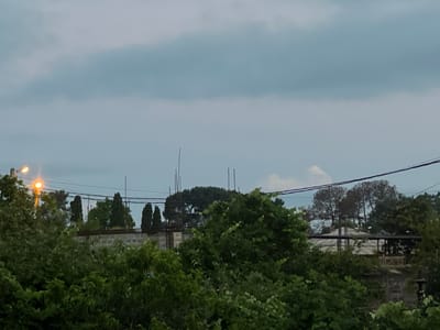 The image depicts an outdoor scene with lush green foliage in the foreground, obscuring much of the background. Above the trees, a hazy sky is visible with some diffused clouds. A street light with an orange glow is present on the left side of the frame, casting a light on the surrounding vegetation. In the midground, there is a low-rise building or wall constructed of what appears to be concrete blocks. Behind this structure, several tall, slender trees and what looks like a larger, denser tree are visible. Power lines crisscross the sky above the trees. In the far background, a partially visible building with a roof structure, possibly a greenhouse or conservatory with a pale roof, can be seen. The overall impression is one of a residential or semi-urban area with significant vegetation. The scene appears to be captured in Ochamchyra, Abkhazia, given the prompt's context.
