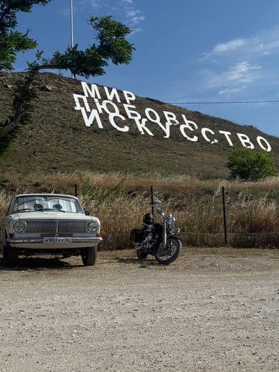 In the city of Sudak, Ukraine, a white vintage car with animated eyes on its windshield is parked on a gravel surface. To the right of the car, a black motorcycle with a helmet on its seat is also parked. Behind the vehicles, a hill is visible with large white lettering spelling out "МИР ЛЮБОВЬ ИСКУССТВО" (Peace Love Art) in Cyrillic. The sky is blue with scattered clouds. The environment appears dry and somewhat arid, with sparse vegetation on the hillside and dry grasses in the foreground. A fence runs behind the parked vehicles.