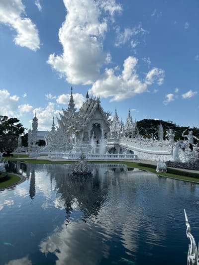 The scene depicts Wat Rong Khun, known as the White Temple, located in Ban Rong Khun, Chiang Rai province, Thailand. A highly ornate, multi-tiered white temple structure, adorned with intricate reflective sculptural details and spires, stands prominently under a clear blue sky with scattered white cumulus clouds. The temple is partially reflected in a dark blue, rippled pond that contains several visible fish and a white sculpted centerpiece. A white, intricately designed bridge, featuring various mythological figures, connects the temple to the foreground. Multiple human figures are visible on this bridge, appearing to be visitors. A fountain operates at the base of the main temple edifice. Green grass borders the pond and extends around parts of the temple complex, with dense green foliage visible in the background on both sides.