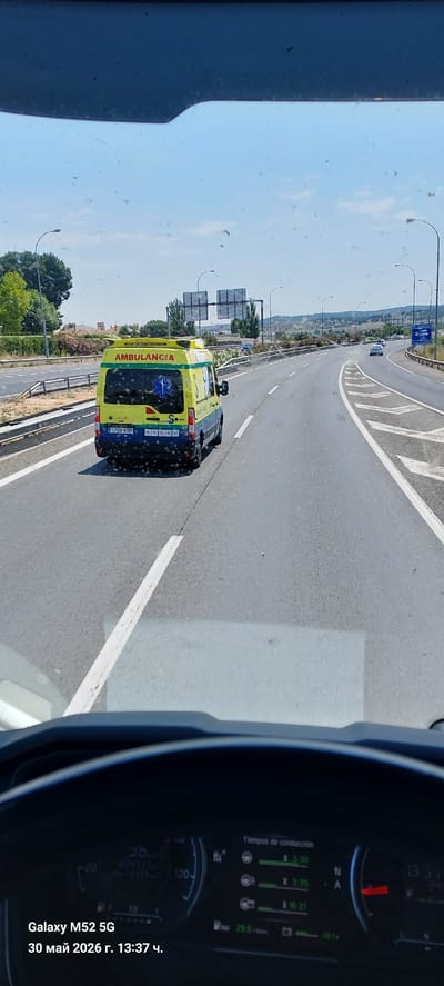 The image, taken from the interior of a vehicle, shows a view of a highway in Toledo, Spain. An ambulance, identified by the word "AMBULANCIA" and blue markings, is driving in the adjacent lane. The road is asphalt, marked with white lane dividers and directional arrows. The foreground includes a portion of the vehicle's dashboard and windshield. The date is 30 Mai 2026 and the time is 13:37. In the background, there are roadside structures, trees, and signage.