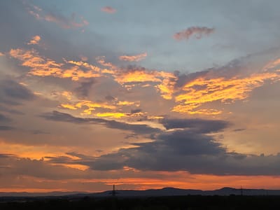 This is an outdoor, daytime, horizontal, eye-level, medium shot photograph of a sunset sky over Freiburg im Breisgau, Germany. The sky is filled with a variety of clouds, ranging from wispy pink and orange clouds to darker, more dramatic grey clouds. The sunset hues create a warm and vibrant atmosphere. In the foreground, a silhouetted line of trees and hills stretches across the bottom of the image. Two power pylons are visible, one to the left and one to the right of center, against the dusky landscape. Several small, dark birds are scattered throughout the sky, adding a sense of scale and movement. The overall scene evokes a sense of natural beauty and the end of a day.