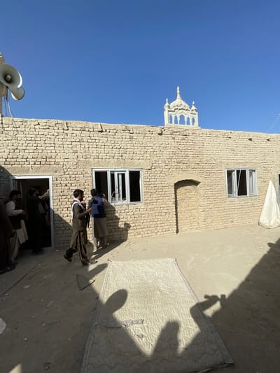 This image depicts an outdoor scene in Spin Boldak, Afghanistan, under a clear blue sky. The primary structure is a single-story building constructed from light-colored bricks, possibly mud-brick or adobe. The building facade features an open rectangular doorway on the far left, a window with white frames to its right (some panes appear missing), a sealed or bricked-up arched doorway in the center, and another white-framed window further right. On the rooftop, to the left, two large, white horn-shaped loudspeakers with visible wiring are mounted. To the right on the rooftop, a white, ornate architectural finial or decorative element is silhouetted against the sky.
Several men are present in the scene. A group of men, dressed in traditional attire, are visible gathered within and around the open doorway on the far left. In the mid-ground, a man in a brown traditional outfit and a dark head covering walks towards the right. A white scarf is draped over his left shoulder, and a dark, long firearm-like object is slung diagonally across his back. Adjacent to him, a second man, wearing a dark blue top and light-colored traditional trousers, stands near the left window, facing it, potentially engaged in an activity related to the window or the building's interior.
The ground is composed of exposed dirt or sand. In the foreground, a large, soiled, light-colored quilted mattress with a distinct pattern lies flat. Strong shadows of multiple individuals, including the photographer, are cast across the mattress and the ground, indicating direct sunlight from the upper left. The scene suggests ongoing activity or a gathering at the brick structure.