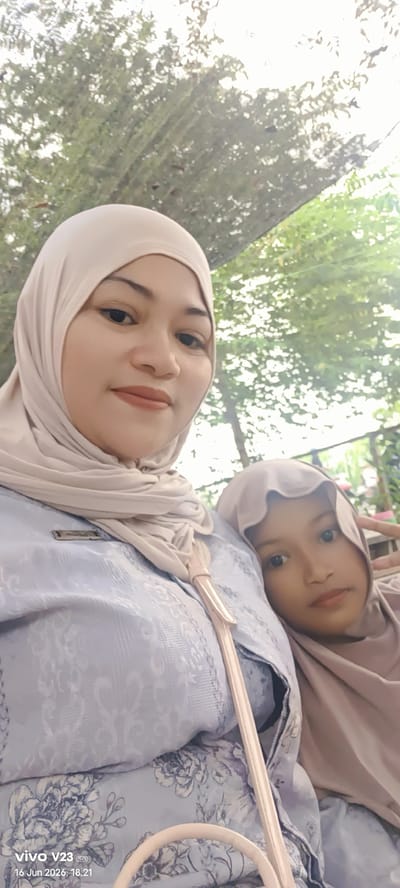 In Glokaron, Indonesia, a selfie is captured outdoors, featuring two individuals. The primary subject, an adult female wearing a beige hijab and a light blue patterned blouse, is positioned in the foreground, looking towards the camera with a slight smile. To her right, partially obscured and leaning against her, is a young girl also wearing a beige hijab. The background consists of lush green foliage and dappled sunlight, suggesting a natural, possibly park-like setting within the city. The lower portion of the image displays a watermark indicating a "vivo V23" phone and the date "16 Jun 2026, 18.21".