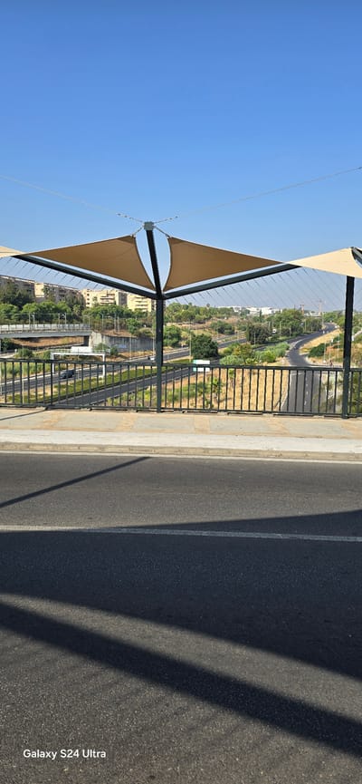 This image captures a view from a pedestrian bridge in San Juan de Aznalfarache, Spain, overlooking a highway. A modern canopy made of taut fabric supported by metal poles provides shade over a section of the bridge. The canopy's fabric is a light brown or tan color, stretched between a central support and outer points. Below the bridge, a multi-lane highway curves through a landscape dotted with trees and buildings. In the distance, several apartment buildings are visible, suggesting a suburban or urban setting. The sky is clear and blue, indicating a sunny day. The foreground of the image shows the asphalt surface of the road, with shadows from the bridge structure cast across it. The overall scene depicts an urban infrastructure element integrated into a natural and built environment.