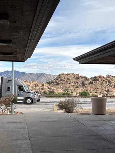 The image shows a gray semi-truck parked on the side of a road, with a mountainous landscape in the background. The truck is facing left, and its side is visible. The landscape consists of rocky hills with sparse vegetation. The foreground shows a concrete walkway and a large, cylindrical trash receptacle. The scene appears to be in an outdoor rest stop or parking area, likely in a desert or semi-arid region. There are two overhanging structures with wood-paneled ceilings and lighting fixtures, suggesting a covered area for vehicles or people. The sky is partly cloudy. The image is captured from a slightly elevated, ground-level perspective. The location is identified as Dragoon, United States.