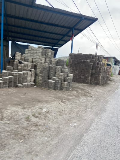 The image shows a pile of concrete bricks stacked on a dirt road in Kecamatan Margaasih, Indonesia. The bricks are arranged in a neat, organized manner, with some bricks stacked higher than others. There is a blue metal awning above the bricks, and a white truck is partially visible in the background. The road is made of asphalt and is relatively narrow, with a dirt shoulder on one side. There are no people in the image.