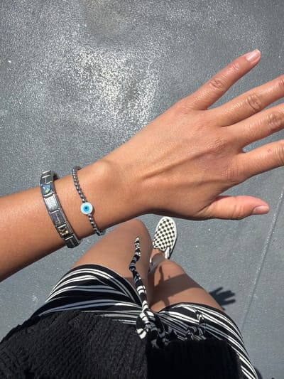 In Point Roberts, United States, a close-up shot captures a person's arm and hand from a first-person perspective. The individual is wearing two bracelets on their left wrist. The first bracelet, closer to the hand, is a silver chain with small, dark beads and a central charm resembling an evil eye with a white iris and blue pupil. Adjacent to this, on the forearm, is a silver link bracelet adorned with various charms, including one with a colorful image and others with text or geometric designs. The person's hand is open, fingers extended, and positioned in the foreground. Their lower body is also visible, showing a black top and a striped skirt with alternating black and white lines. A black and white checkered slip-on shoe is visible on their right foot. The ground beneath them appears to be a textured, gray surface.