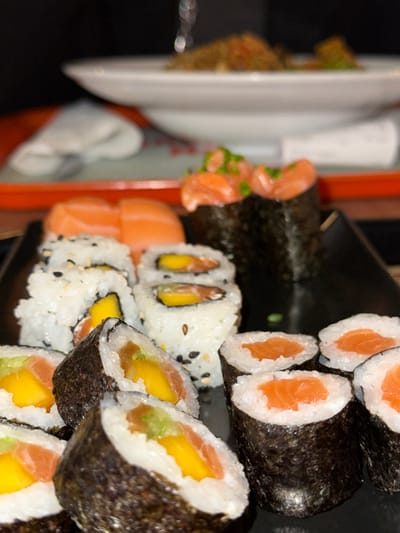 A close-up shows a stack of sushi rolls. The top layer features salmon rolls wrapped in seaweed. Below, there are white rice rolls with black sesame seeds, filled with mango and salmon. Orange salmon pieces are also visible beside these. Plates are visible in the background with a possible dish of salmon topped with herbs.