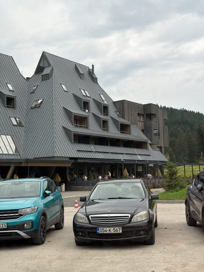 This media file depicts an outdoor scene in a mountainous area, likely in Žunovnica, Bosnia and Herzegovina. The primary subjects are a modern, angular building with a dark gray, metal roof and wooden accents, and two cars parked in front of it. The building appears to be a hotel or a similar establishment, with multiple levels and numerous windows, some of which are recessed or angled.

The activity taking place is not explicit, but the presence of parked cars and some people visible through the building's glass facade suggests a normal operating day for the establishment. A black Mercedes-Benz is parked closest to the building, displaying a license plate that reads "054-K-567". In front of the Mercedes is a turquoise Volkswagen T-Cross, with a partial license plate "115". A small traffic cone with orange and white stripes is positioned between the two cars.

The weather appears to be overcast with a cloudy sky, suggesting it might be morning or late afternoon. The background features a dense, dark green forest covering a steep hillside, typical of a mountainous environment. The overall mood is calm and serene, with no indication of any particular event or strong emotions. The architecture of the building is a notable detail, standing out with its unconventional design against the natural landscape.