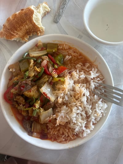 The image displays a meal served on a white tablecloth. The primary subject is a white bowl containing rice and a stir-fry of meat and vegetables, which include green beans, red peppers, and cabbage. A fork rests on the right side of the bowl, partially submerged in the rice. To the left of the bowl is a piece of torn flatbread. In the upper right corner, a white cup filled with a clear liquid is visible. The overall scene suggests a casual dining setting. The provided context indicates this scene is located in the city of Kara-Suu, Kyrgyzstan.