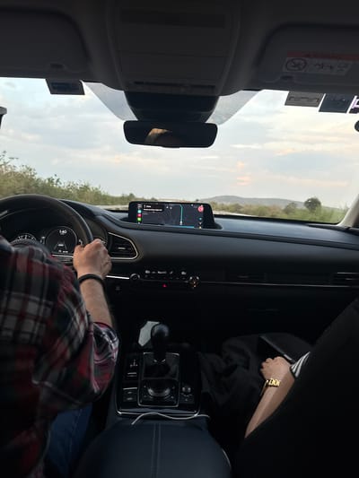 The image captures a scene from inside a car, likely a Mazda, during a drive near San Juan Tejupa, Mexico. The driver, wearing a red and black plaid shirt and a black bracelet, has both hands on the steering wheel. The dashboard display indicates a navigation system pointing to "Caseta de Cobro Siglo XXI" approximately 9 kilometers ahead. A passenger can be partially seen on the right, with a glimpse of a gold watch on their wrist. The car appears to be driving on a road surrounded by scrub vegetation under a sky partially covered with light clouds. The sun is casting long shadows. The speedometer reads near 140, which is most likely 140 km/hr.