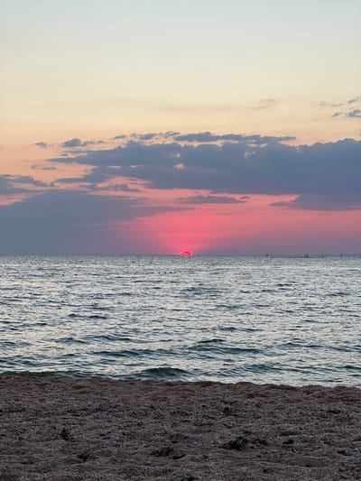The image depicts a sunset over a body of water, likely the Black Sea, near the village of Chelidynove, Ukraine. The sky transitions from a pale yellow at the top to hues of pink and purple closer to the horizon. Dark, scattered clouds are present in the upper half of the sky. The sun, a vibrant pink orb, is partially visible just above the horizon line. The sea is calm, with gentle waves creating a textured surface. In the distance, several vertical poles are visible, suggesting structures or markers in the water. The foreground of the image shows a beach covered in small shells and pebbles. No people or animals are visible in the frame.