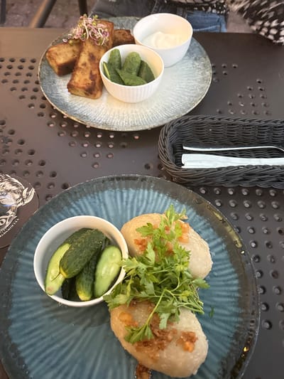 The image depicts a close-up view of two plates of food on a dark, perforated table, likely outdoors at a restaurant in Hajnówka, Poland. The plate in the foreground contains two potato dumplings garnished with fried onions and fresh parsley, served with a small bowl of pickles. The plate in the background shows several rectangular slices of fried bread, a small bowl of pickles, and a bowl of white sauce. A black woven basket holding cutlery is visible to the right. The lighting suggests it is daytime.