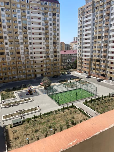 This is a high-angle, medium shot of a residential complex in Sumgayit, Azerbaijan, likely taken from a balcony. Two tall, modern apartment buildings with tan and white facades dominate the left and right sides of the frame. Numerous windows and balconies punctuate the buildings' exteriors. In the foreground, a red-orange, textured railing is visible. Between the apartment buildings, a communal outdoor area features a children's playground with colorful equipment, a paved courtyard, and a fenced-in green sports court, possibly for futsal or volleyball. Several trees and landscaped garden beds are scattered throughout the area. A few people are present, including children on the playground and adults walking in the courtyard and near the garden. Several cars are parked on the asphalt road bordering the complex. In the background, other residential buildings with red-tiled roofs are visible, suggesting a densely populated urban environment. The sky is clear and blue, indicating a sunny day.