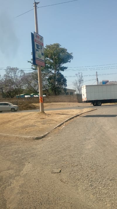A daytime street scene features a roadside sign attached to a utility pole. The sign reads "AM-X Body Parts" with an image. A light-colored sedan is partially visible on the left. Behind the pole is dry grass and a low brick wall. A white box truck is moving to the right. The sky is blue. The road in the foreground is cracked and worn.
