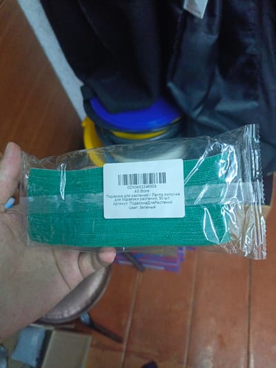 The image shows a close-up view of a person's hand holding a package of green plant ties. The package has a white label with text in Russian. The text indicates that the product is a Velcro tie for plants, with "50 pieces" and the color "Green". The barcode is also visible. The background appears to be indoors, with some blurry objects and a wooden surface. The location in the city of Ухманы, Russia is not discernible from the image itself.