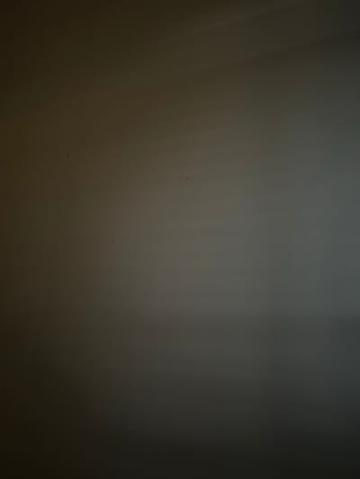This media file is a photograph of a wall, likely indoors. The wall has a textured surface and is dimly lit, with a gradient of light and shadow that suggests a light source off to the left. There are several small, dark specks visible on the surface of the wall, which could be dust or imperfections. The overall impression is that of a plain, unadorned wall in a neutral or subdued environment. There are no discernible objects, people, or activities taking place. The scene is devoid of any specific environmental cues that would indicate the time of day, weather, or a precise location, beyond the general context of Ravenna, Italy. No text is visible in the image.