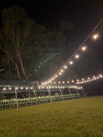 This media file captures an outdoor evening scene at Mururiini, Kenya, illuminated by numerous string lights. The image, originally rotated 90 degrees clockwise, shows a dark night sky at the top. On the left, lush green trees are visible against the dark background, with the warm glow of the lights reflecting subtly off their leaves.
Multiple strands of warm-toned string lights stretch across the upper and middle portions of the frame, creating a festive ambiance. Below these lights, a structure, possibly a tent or covered area for an event, runs horizontally across the mid-ground. Attached to this structure are rows of light-colored chairs, arranged neatly as if for an audience, suggesting an event setup. A speaker is visibly mounted on a pole within this structured area. To the right, a well-maintained grassy field or lawn extends, illuminated softly by the overhead lights.
The scene appears to be set for a public gathering, concert, or celebration. The time of day is clearly night, and the weather seems clear. No people are visible, indicating either the event has not yet started, has ended, or the focus is on the setup itself. There is no visible text.