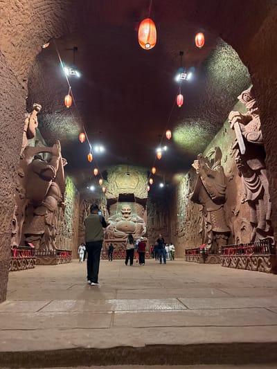 The image shows the interior of the Dazu Rock Carvings in Shizhong, China. The scene is a cave-like structure with carved figures along the walls. Red lanterns illuminate the space, suspended from the ceiling. Several people are present, observing and photographing the carvings. The walls and floor are primarily stone, with intricate carvings decorating the walls. Some carvings include reclining figures.