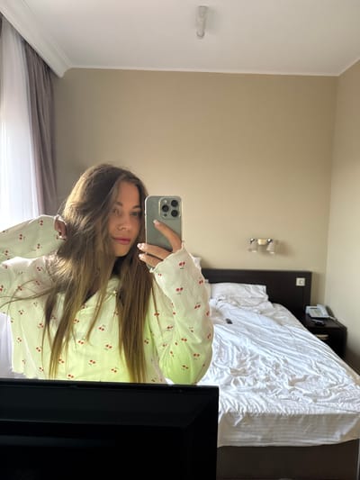 A young woman is taking a selfie in a bedroom. The bedroom contains a bed with white linens, a dark headboard, a lamp, and a television. The woman is wearing a white, long-sleeved shirt with a cherry pattern. It is not possible to verify if the location is in Sosnovy, Russia from the image alone.