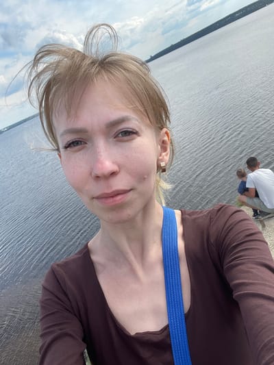 A selfie-style portrait shows a young woman in the foreground, with a vast body of water and distant land in the background. The woman has light-colored hair, which is styled loosely around her face, and light eyes. She wears a dark brown long-sleeved top and a prominent blue strap, possibly from a bag, crosses her shoulder. A small gold-colored earring is visible in her right ear. She looks directly at the camera with a neutral to slightly smiling expression.

In the mid-ground, near the edge of the water, two men and a small child are visible. One man wears a white t-shirt and grey shorts, kneeling or crouching, while the child in a blue t-shirt is close to him. Another man is partially obscured. They appear to be engaged in a quiet activity near the water.

The setting features a large expanse of water, likely a lake or wide river, with subtle ripples on its surface. A strip of land, covered with trees or vegetation, is visible on the distant horizon, separating the water from the sky. The sky above is cloudy, suggesting an overcast or partly cloudy day. The time of day appears to be daytime, with diffused light. The location is specified as Пятый километр, Russia. There is no visible text in the image.