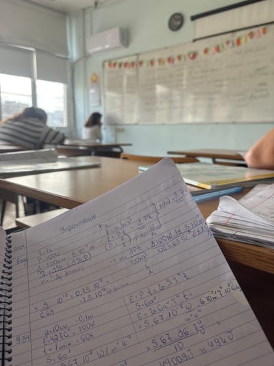 A close-up shot focuses on a notebook page filled with handwritten physics calculations, seemingly related to energy and radiation. The calculations involve variables like power (P), wavelength (λ), temperature (T), and Stefan-Boltzmann constant (σ), suggesting topics such as blackbody radiation or heat transfer.
The notebook sits on a wooden desk, partially obscured by other papers and a pen. In the background, a classroom setting is visible, complete with a whiteboard covered in writing, a radiator, and students at desks. The environment suggests an educational context, possibly a physics class in session.
While the location appears to be a classroom setting, there is no visual information that suggests the image was taken in the city of Trilistnik, Bulgaria.