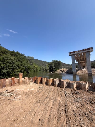 The image depicts a construction site in the city of Kresna, Bulgaria, focusing on a bridge under construction over a river. In the foreground, a section of the riverbank is reinforced with metal sheet piling and wooden posts, with disturbed earth and a coiled rope visible on the muddy ground. The river flows calmly, reflecting the clear blue sky and surrounding lush green hillsides covered in trees. A partially constructed concrete bridge dominates the middle ground, supported by large concrete pillars rising from the water. The bridge deck is incomplete, with exposed rebar at the edges. The overall scene suggests ongoing infrastructure development in a natural, rural setting.