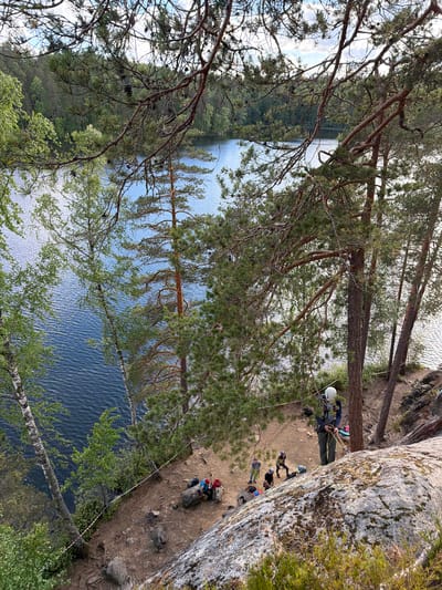 The image depicts an outdoor scene by a body of water, likely a lake, surrounded by lush green trees. In the foreground, a large, moss-covered rock dominates the lower portion of the frame, with a person in climbing gear standing on it, holding ropes. Several other individuals are visible in the background on a dirt path near the water's edge, some sitting on rocks and one appearing to be engaged in an activity with another person. The environment suggests a recreational area with a focus on climbing or outdoor activities. The presence of a lake and dense forest points towards a natural landscape. This scene is not located in the city of Kulikovo, Russia; rather, it is a natural, forested area.