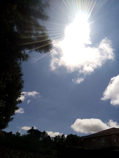 This image, captured in Nachu ward, Kenya, depicts a bright outdoor scene dominated by a clear blue sky with scattered white cumulus clouds. The sun is prominently featured in the upper right quadrant, radiating intense sunbeams. The left side and upper left corner of the frame are filled with the dark, silhouetted foliage of a large tree, heavily underexposed by the backlight. A distinct dark treeline forms a horizontal band across the lower middle section of the image. In the lower right corner, a portion of a building with a reddish-brown tiled roof is partially visible, also appearing largely silhouetted. No individuals or specific activities are present.