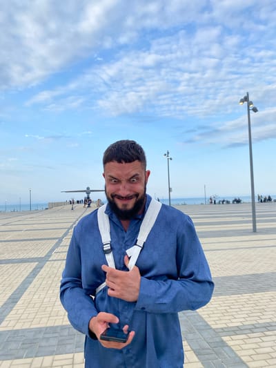 A man with a beard and short dark hair is standing outdoors, looking at the camera with a mischievous expression. He is wearing a blue button-down shirt with a repeating pattern of the Gucci logo. A white strap goes over his left shoulder and across his chest. He is holding a black smartphone in his right hand. The background shows a paved promenade with light and dark gray tiles, a lamppost on the right, and a body of water in the distance. There is a large, abstract sculpture in the middle ground, with several people visible around it. The sky is blue with scattered white clouds. The location is identified as Arablyar, Russia.