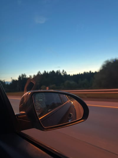 A view from inside a car at dusk, looking out the passenger side window. The sky is a gradient of deep blue to lighter hues near the horizon, with a faint, wispy cloud. A dense line of dark green pine trees forms the background, with a lighter colored strip, possibly a road or fence, running horizontally across the middle ground.
The side mirror of the car is prominently featured, reflecting a distorted image of the car's interior, including a person holding a smartphone. The car's interior is dimly lit, with warm, orange light reflecting off surfaces. The road visible outside the car is also illuminated with orange light, suggesting streetlights or the setting sun. The overall impression is of a serene, twilight drive on a highway, possibly on the outskirts of a forested area.
The image does not contain enough visual information to determine a specific location within Khrustal'nyy, Russia.