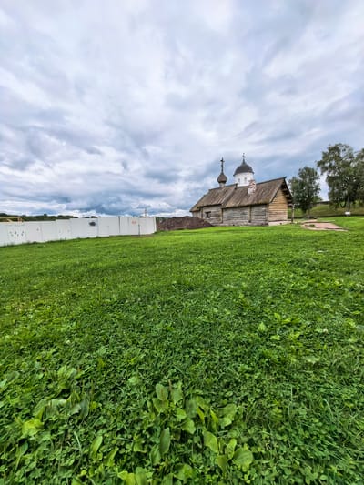 An outdoor, full shot captures a traditional wooden church with two onion domes and a cross atop the higher dome. The church is constructed of rough-hewn logs and has a wooden shingle roof. To the left of the church, a white temporary construction fence stretches across the frame, with some construction materials and a pile of dark soil visible beyond it. The foreground is dominated by a lush green grassy field. In the background, several trees are visible, and the sky is overcast with dramatic, grey clouds. The scene appears to be in Staraya Ladoga, Russia, likely a historical or cultural site. There are no people present in the image.