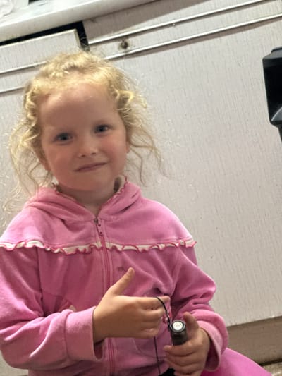 A young girl with blonde curly hair is shown. She is wearing a pink hooded jacket with white ruffled trim. Her right hand is holding a small, cylindrical object. The girl is looking towards the camera with a slight smile. The background appears to be a light-colored, textured surface, possibly a wall or door. The scene is indoors, with the lighting suggesting it could be daytime. There is no visible text in the image.
