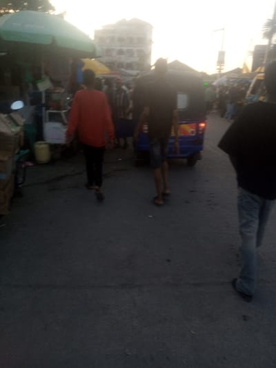 This image depicts a bustling outdoor street or market scene in Mtopanga ward, Kenya, observed under soft, diffused light consistent with late afternoon or early evening. The ground is an asphalt surface. In the immediate foreground, the backs of three individuals are visible, walking away from the camera. The person on the far right wears a dark top and light-colored jeans. The central figure wears a dark short-sleeved top and dark shorts. The person on the left wears a red long-sleeved top and dark trousers.
A blue three-wheeled auto-rickshaw, or tuk-tuk, with active rear lighting, is positioned in the central midground, also facing away from the camera. To the left of the scene, a market stall features a large green canopy, with a yellow jerrycan and stacked cardboard boxes visible on the ground nearby. A motorcycle's front section is partially visible adjacent to this stall. Further into the background, indistinct figures populate the area, alongside additional market structures, including a yellow canopy. A multi-story building is silhouetted against a bright, pale sky in the distant background. The scene indicates active pedestrian and limited vehicular movement within a commercial thoroughfare.