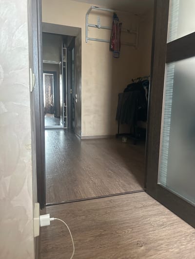 This is a downward-angle, interior shot of a hallway and what appears to be a doorway or entrance. The scene is illuminated by artificial light, and the time of day and weather are not discernible.
In the foreground, a white electrical outlet with a white charging brick and a white cable plugged into it is visible. The outlet is situated on a textured, possibly marbled surface. This charging setup is positioned on the edge of a dark wooden structure, which forms the frame of a view into the hallway.
The hallway itself features light-colored walls and a wooden-patterned floor. A metal drying rack with some items on it is mounted on the wall. Further down the hallway, a doorway opens into what appears to be a more dimly lit area, possibly another room or apartment entrance. This area contains shelves with various items, including what looks like clothing, and a framed picture on the wall. A glimpse into another section of the hallway shows a patterned carpet.
The overall impression is one of a functional, domestic hallway. The scene is static, with no people or active events taking place. The location cues are limited to the interior setting, and the colors are muted, consisting primarily of browns, beiges, and whites. No visible text is present in the image.