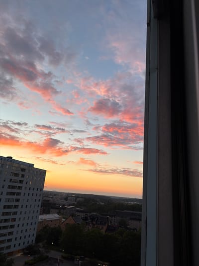 A vertical, high-angle, medium shot captures a vibrant sunset over the city of Solna, Sweden, as viewed from a building. In the foreground, a portion of a modern, light-colored apartment building occupies the left side of the frame, its numerous rectangular windows creating a geometric pattern. To the right, the dark, vertical lines of a window frame divide the image, offering a glimpse of the exterior.

The sky is a spectacle of color, transitioning from a pale blue at the top to a warm orange and yellow gradient near the horizon. Wispy clouds, tinged with shades of pink and orange, are scattered across the sky, illuminated by the setting sun.

Below the sky, the cityscape of Solna unfolds. A dense residential area with a mix of apartment buildings and houses is visible, interspersed with lush green trees. The buildings appear to be a mixture of older and newer constructions. The overall impression is of a suburban or city outskirts landscape bathed in the soft, warm light of dusk. No people are visible in the frame.