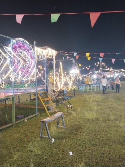 The image shows a nighttime scene in Menjing, Indonesia, likely a local carnival or fair.
Foreground: The foreground is dominated by tall, slightly overgrown grass. There's a weathered wooden sawhorse, and a set of metal stairs leading to a fenced-in platform.
Midground: In the midground, several amusement rides are visible. These appear to be simple carnival attractions illuminated with strings of white lights. People walk in the distance between rides.
Background: Overhead, colored flags or bunting are strung up, adding to the festive atmosphere. Above the lights and decorations is a dark night sky.