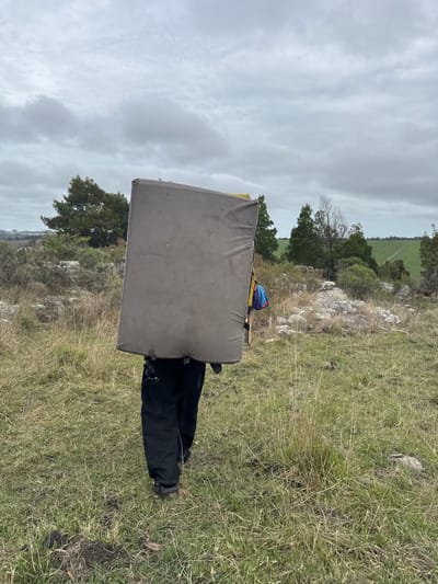 An individual is depicted from behind, traversing an open, grassy landscape in Sierra de los Padres, Argentina. The person is wearing black pants and dark footwear. A large, rectangular, grey-brown bouldering crash pad is strapped to their back, obscuring their upper body and head. A small, multicolored item is visible peeking from behind the right side of the crash pad. The individual is walking away from the camera on uneven ground covered with dry and green grass and scattered rocks. In the middle ground, various trees and bushes are present, with a flat, green agricultural field discernible in the distant background on the right. The sky is overcast with dense grey clouds.