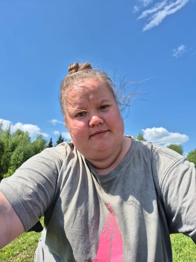 A medium-shot, ground-level, rotated selfie captures a fair-skinned woman against a backdrop of blue sky with scattered white clouds, and green treetops. The woman is wearing a grey t-shirt with a faded pink design. Her hair is pulled back in a bun. The location is outdoors, suggested by the natural elements in the scene. There is no indication of the location being in the city of Дойная, Russia.
