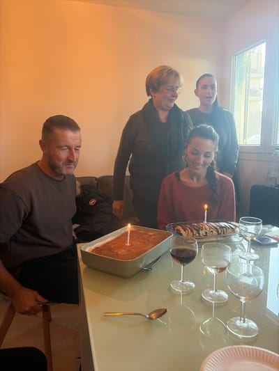 A group of four individuals is gathered around a table in what appears to be an indoor residential setting in Cagnes-sur-Mer, France. A man in a dark t-shirt is seated on the left, looking towards the center-right with a slight smile. A woman with dark braided hair and a reddish-brown top is seated on the right, smiling broadly and looking down at the table. Behind her stands an older woman with blonde hair and glasses, wearing a dark top and scarf, smiling and looking towards the seated woman. To her right, another younger woman with dark hair stands, mouth slightly ajar, looking in the same direction.
The light-colored, reflective table in the foreground holds two desserts with lit white candles: a rectangular dish containing a brown, layered dessert, and a clear glass dish with a rolled pastry topped with chocolate drizzle. Three wine glasses are visible; one contains red liquid, while the other two are empty. A single silver spoon rests on the table. A white paper plate is partially visible in the lower right corner.
Behind the seated man, a dark sofa is partially visible, with a dark jacket featuring a red circular logo draped over it. A large window on the right side of the frame reveals the exterior of multi-story residential buildings, consistent with an urban environment in Cagnes-sur-Mer. The walls are plain and light-colored.