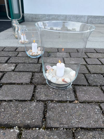 Two glass hurricane lanterns with white pillar candles and sand with seashells are placed on a paved outdoor surface. The lanterns are of different sizes, with the larger one behind and to the side of the smaller one. Both candles are lit, with visible flames. The larger lantern contains more sand and seashells than the smaller one. In the background, there is a glimpse of a building with a green door and metal railing, suggesting an entrance. The paving stones are dark gray and appear to be made of concrete with small pebbles. The scene is outdoors, and the lighting suggests it could be daytime. The overall impression is one of outdoor decoration, possibly for an event or to enhance a patio area. There is no visible text in the image. The location context is Ufhusen, Switzerland.